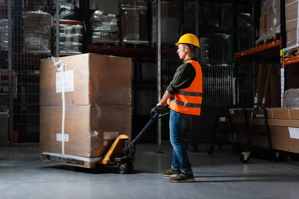Strong middle aged warehouse worker in hard hat and safety vest transporting pallet with hand truck — Stock Photo