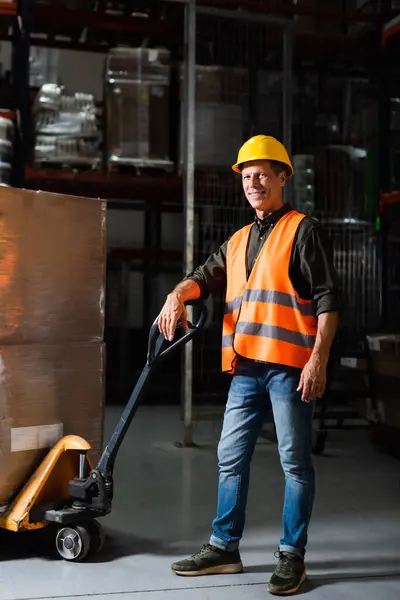 Happy middle aged warehouse worker in safety vest transporting heavy pallet with hand truck — Stock Photo