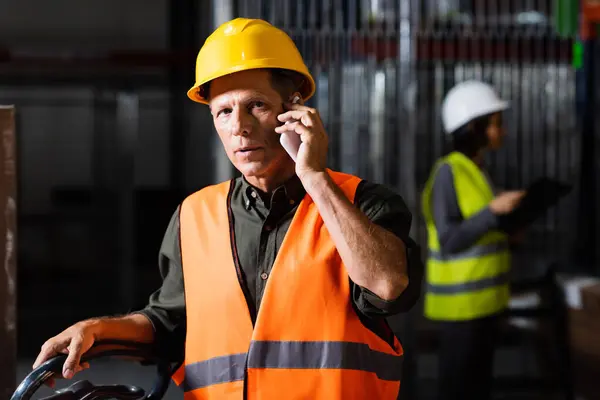 Warehouse supervisor making a phone call on smartphone with a female colleague in the background — Stock Photo