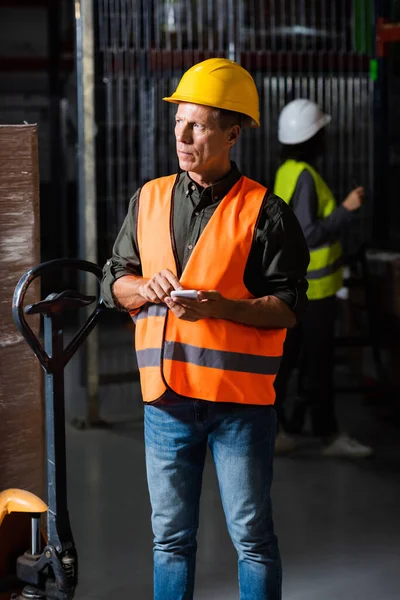 Supervisor de almacén en chaleco de seguridad y casco usando smartphone con empleado en segundo plano - foto de stock