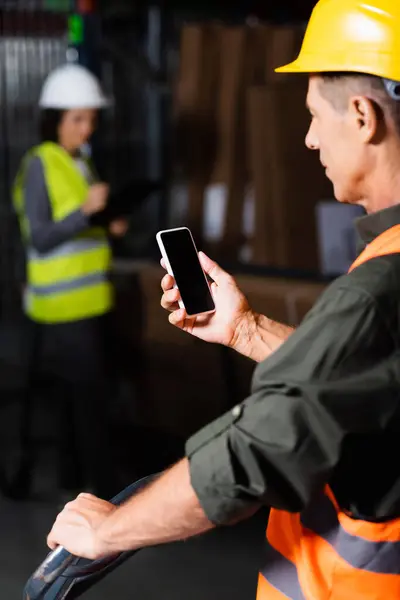Supervisore in giubbotto di sicurezza e casco utilizzando smartphone con dipendente sullo sfondo del magazzino — Foto stock