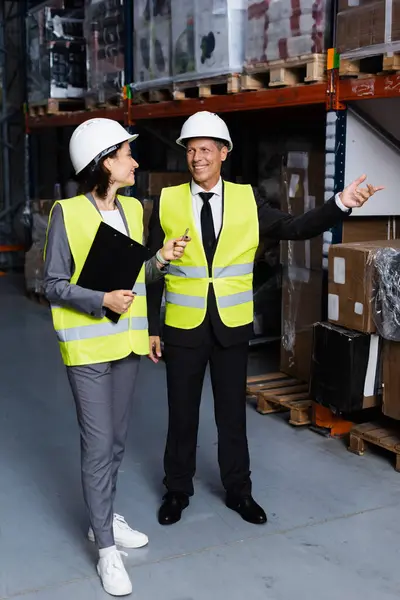 Happy warehouse supervisor instructing female employee in hard hat and safety vest, showing cargo — Stock Photo