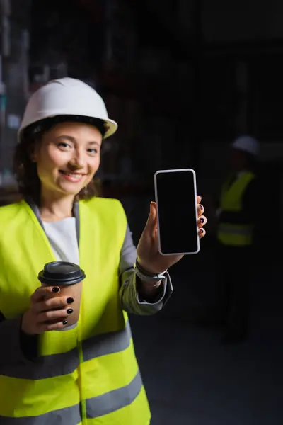 Mulher sorridente em chapéu duro segurando smartphone e copo de papel, feliz funcionário do armazém no coffee break — Fotografia de Stock