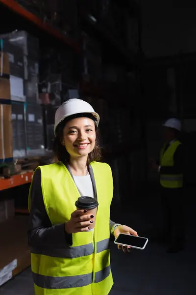 Mulher em chapéu duro segurando smartphone e copo de papel, feliz funcionário do armazém no coffee break — Fotografia de Stock