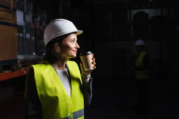 Vista lateral da mulher em chapéu duro segurando copo de papel, feliz funcionário do armazém durante a pausa para o café — Fotografia de Stock