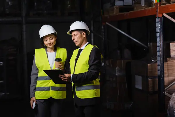Arbeiter mittleren Alters in harter Mütze zeigt einer Mitarbeiterin Klemmbrett mit Kaffee und Smartphone — Stockfoto
