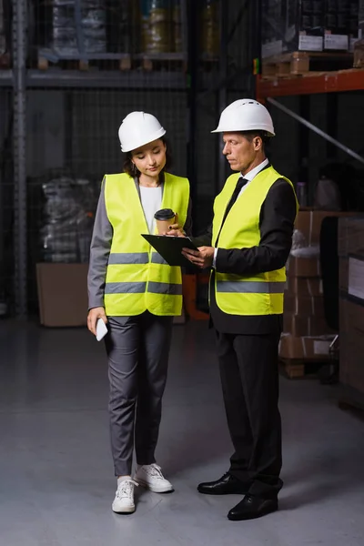 Warehouse supervisor in hard hat showing clipboard to female employee with coffee and smartphone — Stock Photo