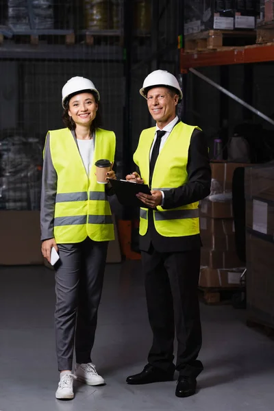 Warehouse supervisor with clipboard and female employee with coffee and smartphone looking at camera — Stock Photo