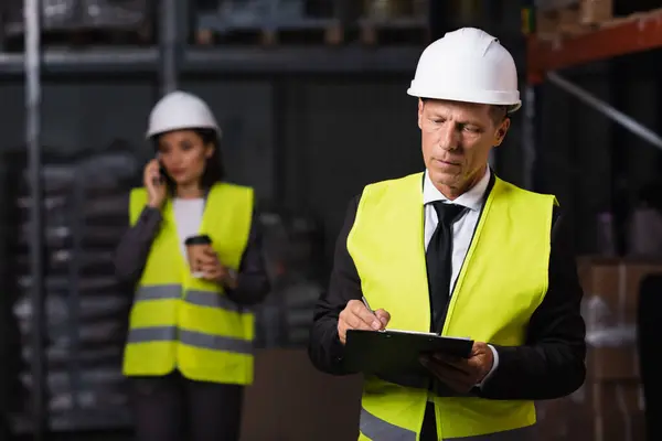 Warehouse supervisor in hard hat writing on clipboard with female employee on blurred background — Stock Photo