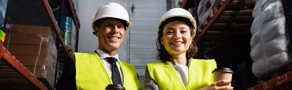 Cheerful logistics team with hard hats and coffee smiling during a break in warehouse, banner — Stock Photo