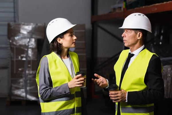 Equipo de logística, mujer en sombrero duro y café charlando con supervisor de mediana edad en el almacén - foto de stock