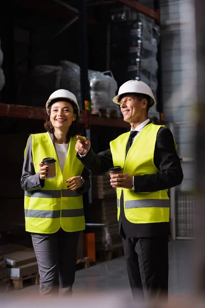 Logistik-Team, glücklicher Vorgesetzter mit Hut und Kaffee, der dem Kollegen im Lager etwas zeigt — Stockfoto