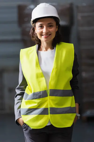 Femme professionnelle heureuse en gilet de sécurité et chapeau dur debout avec les mains dans les poches dans l'entrepôt — Photo de stock
