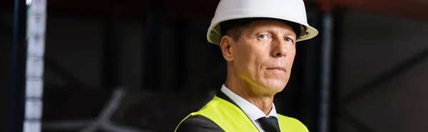 Confident man in safety vest and hard hat looking at camera in warehouse, professional banner — Stock Photo