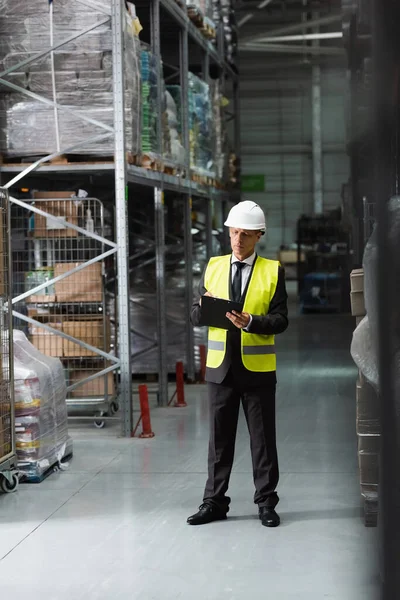 Confident middle aged supervisor in safety vest and hard hat writing on clipboard in warehouse — Stock Photo