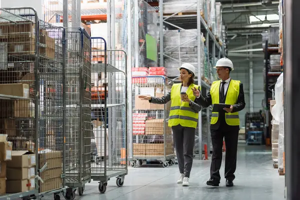 Logistics team with hard hats walking with coffee near inventory while inspecting warehouse — Stock Photo
