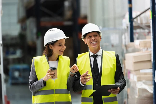 Logistics workers with hard hats walking with coffee near inventory while inspecting warehouse — Stock Photo