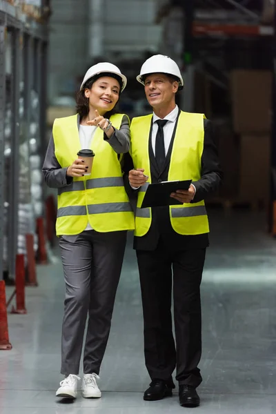 Trabalhadores logísticos felizes com chapéus duros andando com café perto do inventário enquanto inspeciona o armazém — Fotografia de Stock