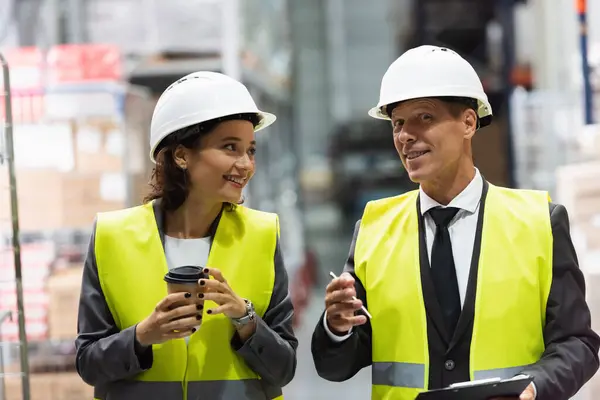Trabajadores logísticos felices en sombreros duros caminando con café cerca del inventario mientras inspeccionan el almacén - foto de stock