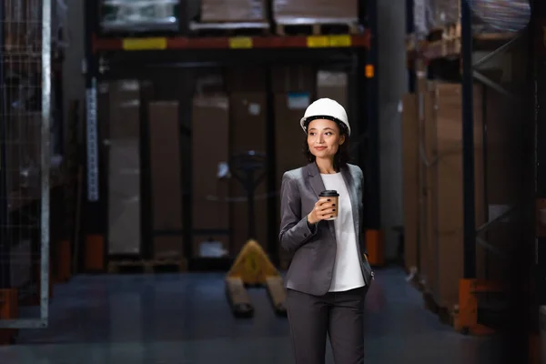 Happy logistics team in hard hats walking with coffee near inventory while inspecting warehouse — Stock Photo