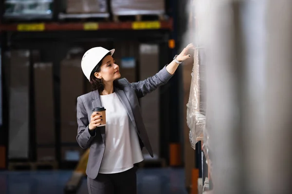 Donna d'affari in giacca e cravatta e cappello in possesso di caffè per andare in magazzino, gestione logistica — Foto stock