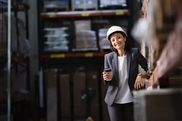 Mujer de negocios en traje y oído sombrero celebración de café para ir e inspeccionado inventario en el almacén - foto de stock