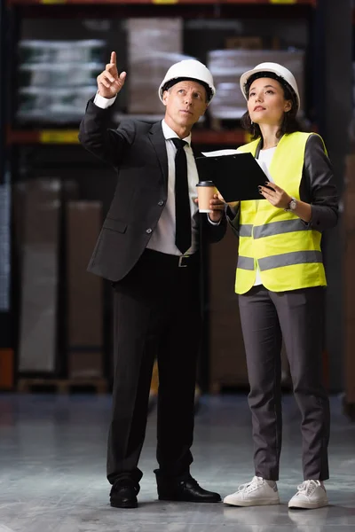 Homem de negócios de terno e chapéu duro planejamento de operações logísticas com funcionária feminina em colete de segurança — Fotografia de Stock