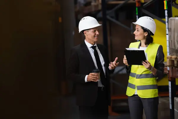 Happy businessman in suit and hard hat discussing logistics operations with female employee — Stock Photo