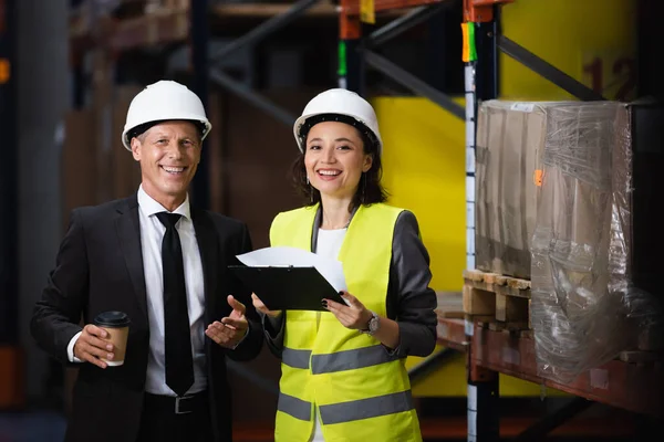 Sonriente hombre de negocios en traje y sombrero duro sosteniendo café cerca de empleada femenina, concepto de logística - foto de stock