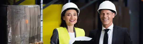 Hombre de negocios feliz en traje y sombrero duro sonriendo cerca de empleada femenina, bandera logística - foto de stock