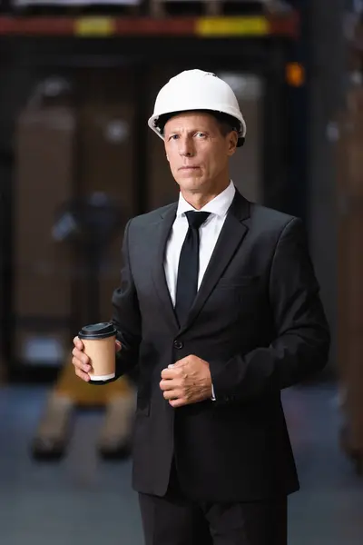 Confident middle aged businessman in hard hat and suit holding coffee in warehouse, headshot — Stock Photo