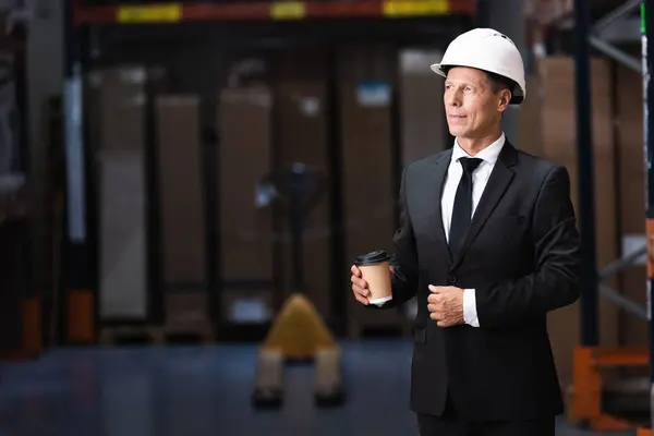 Complacido hombre de negocios de mediana edad en sombrero duro y traje que sostiene el café en el almacén, profesional - foto de stock
