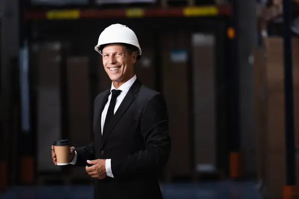 Happy middle aged businessman in hard hat and suit holding coffee in warehouse, professional — Stock Photo