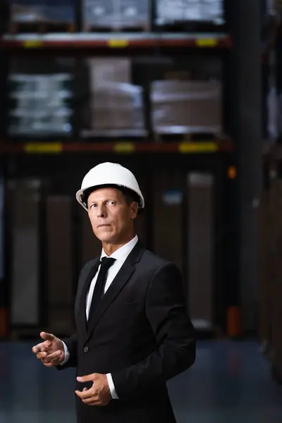 Serious middle aged businessman in hard hat and suit standing warehouse, professional headshot — Stock Photo