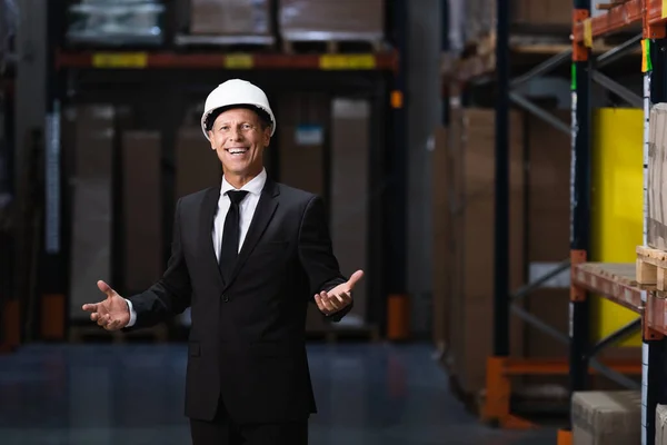 Friendly middle aged businessman in hard hat and suit gesturing in warehouse, professional headshot — Stock Photo