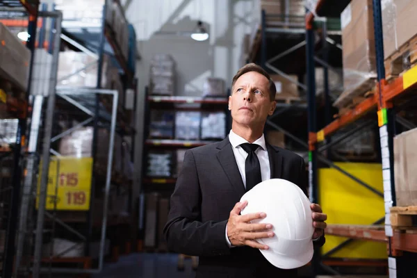 Pensive middle aged businessman in suit holding hard hat in warehouse, professional headshot — Stock Photo