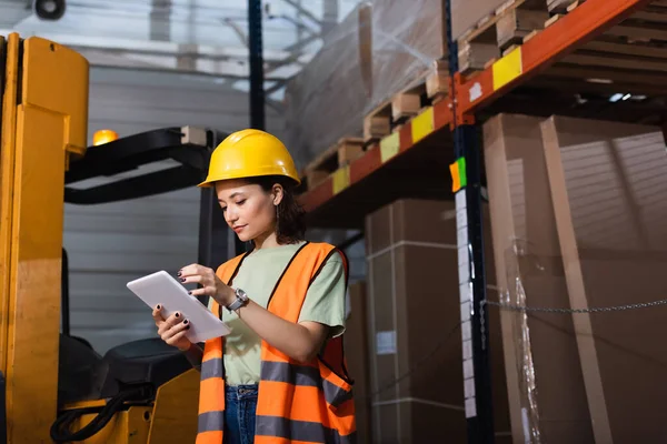 Female warehouse worker in hard hat and safety vest using digital tablet near forklift, cargo — Stock Photo