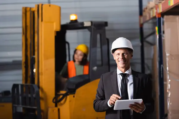 Middle aged warehouse supervisor in suit and hard hat using tablet while smiling at camera — Stock Photo