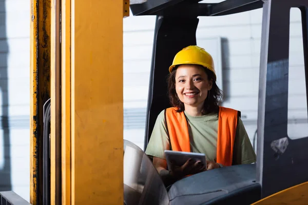 Female forklift operator in hard hat and safety vest smiling and holding tablet in warehouse — Stock Photo