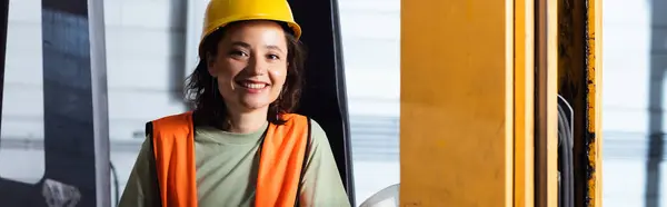 Operador de empilhadeira fêmea em chapéu duro e colete de segurança sorrindo no armazém, bandeira horizontal — Fotografia de Stock