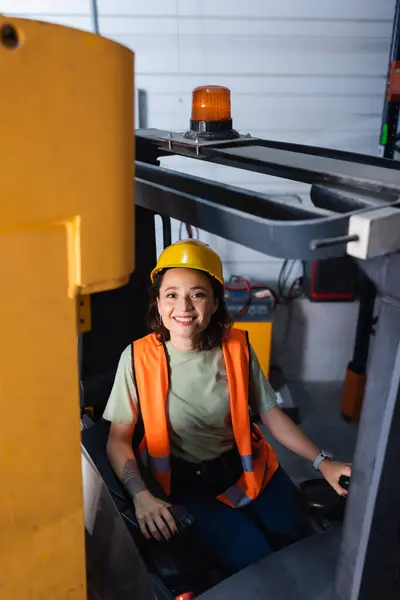Cheerful female forklift operator in hard hat and safety vest looking at camera in warehouse, cargo — Stock Photo