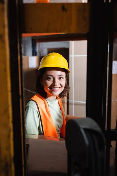 Happy female forklift operator in hard hat and safety vest looking at camera in warehouse, cargo — Stock Photo