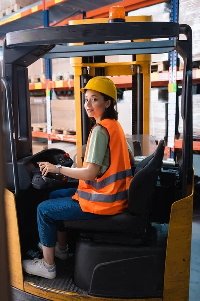 Heureux opérateur de chariot élévateur féminin en casque dur et gilet de sécurité regardant la caméra dans l'entrepôt, la logistique — Photo de stock