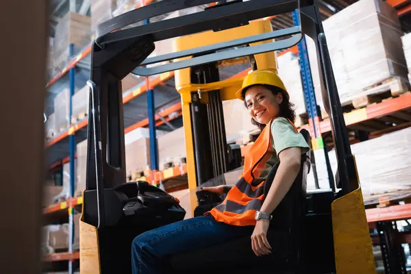 Happy female forklift operator in hard hat and safety vest looking at camera in warehouse, cargo — Stock Photo