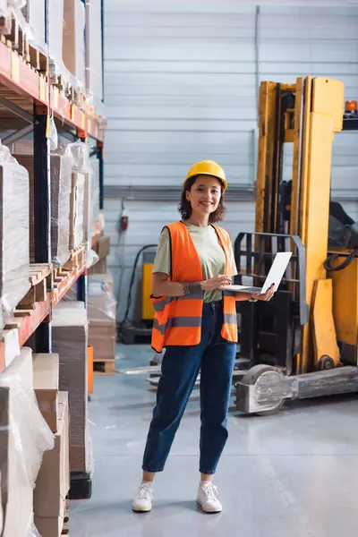 Happy warehouse female worker in hard hat and safety vest using laptop while checking inventory — Stock Photo