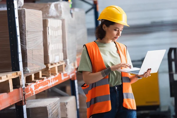 Female worker in hard hat and safety vest using laptop while checking cargo in warehouse, management — Stock Photo