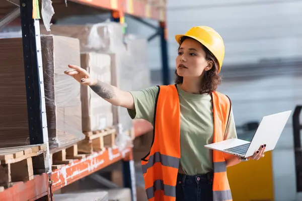 Female worker in hard hat and safety vest using laptop while pointing at cargo in warehouse — Stock Photo