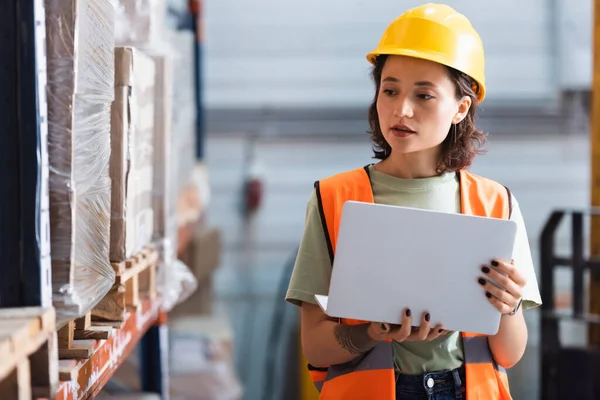 Female worker in hard hat and safety vest using laptop while checking inventory in warehouse — Stock Photo