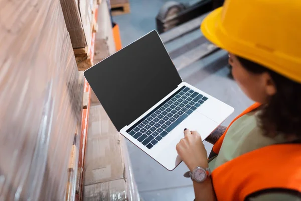 Trabajadora de almacén en sombrero duro y chaleco de seguridad utilizando el ordenador portátil mientras revisa la carga, los gastos generales - foto de stock