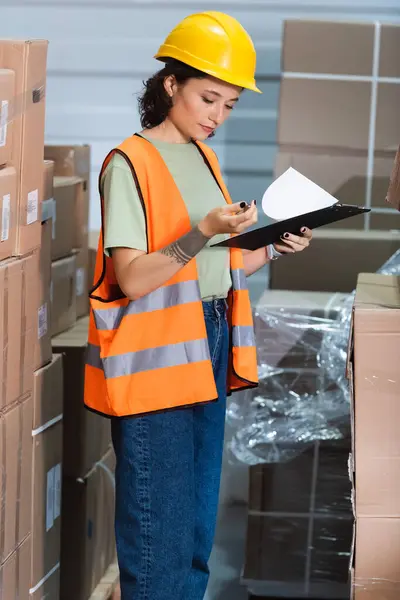 Female warehouse worker in hard hat and safety vest holding clipboard checking inventory — Stock Photo